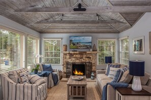 A cozy living room with a stone fireplace, striped sofas, and large windows with blinds.