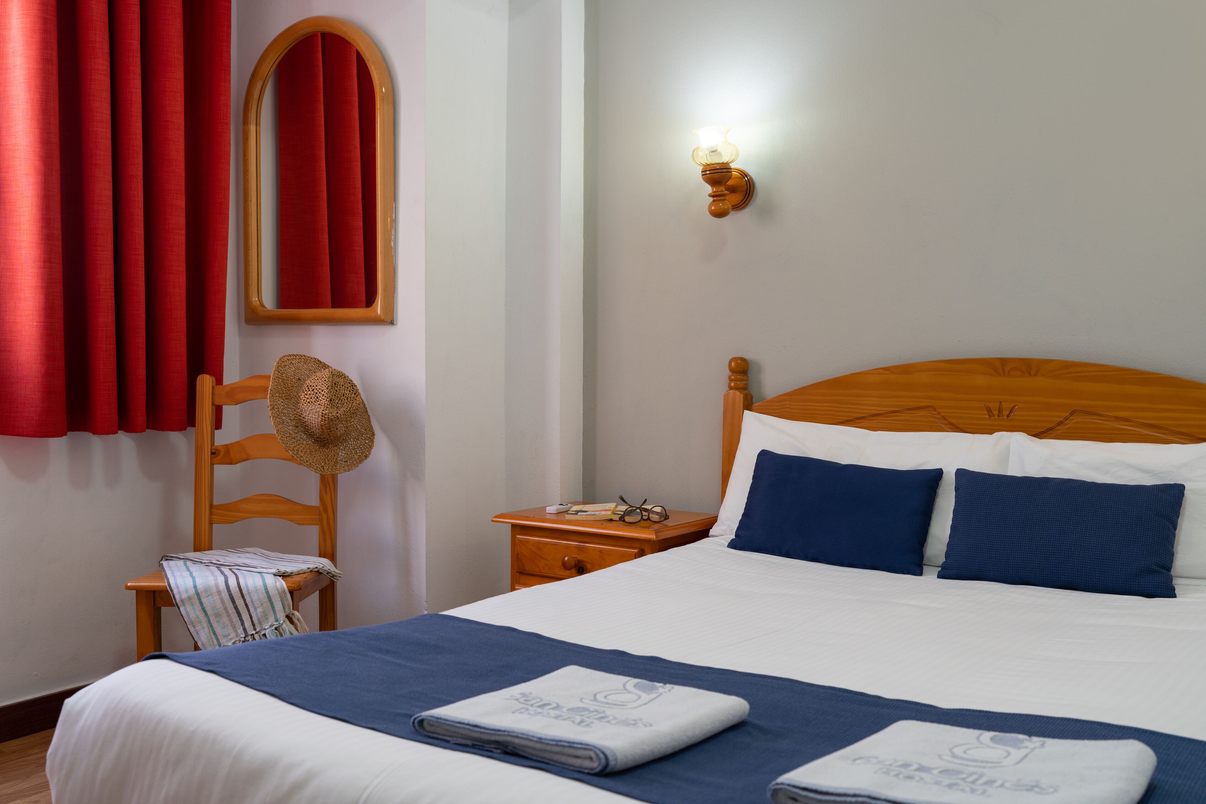 Una habitación de hotel con una cama, una mesita de noche de madera, una silla, un sombrero y una lámpara fijada a la pared.