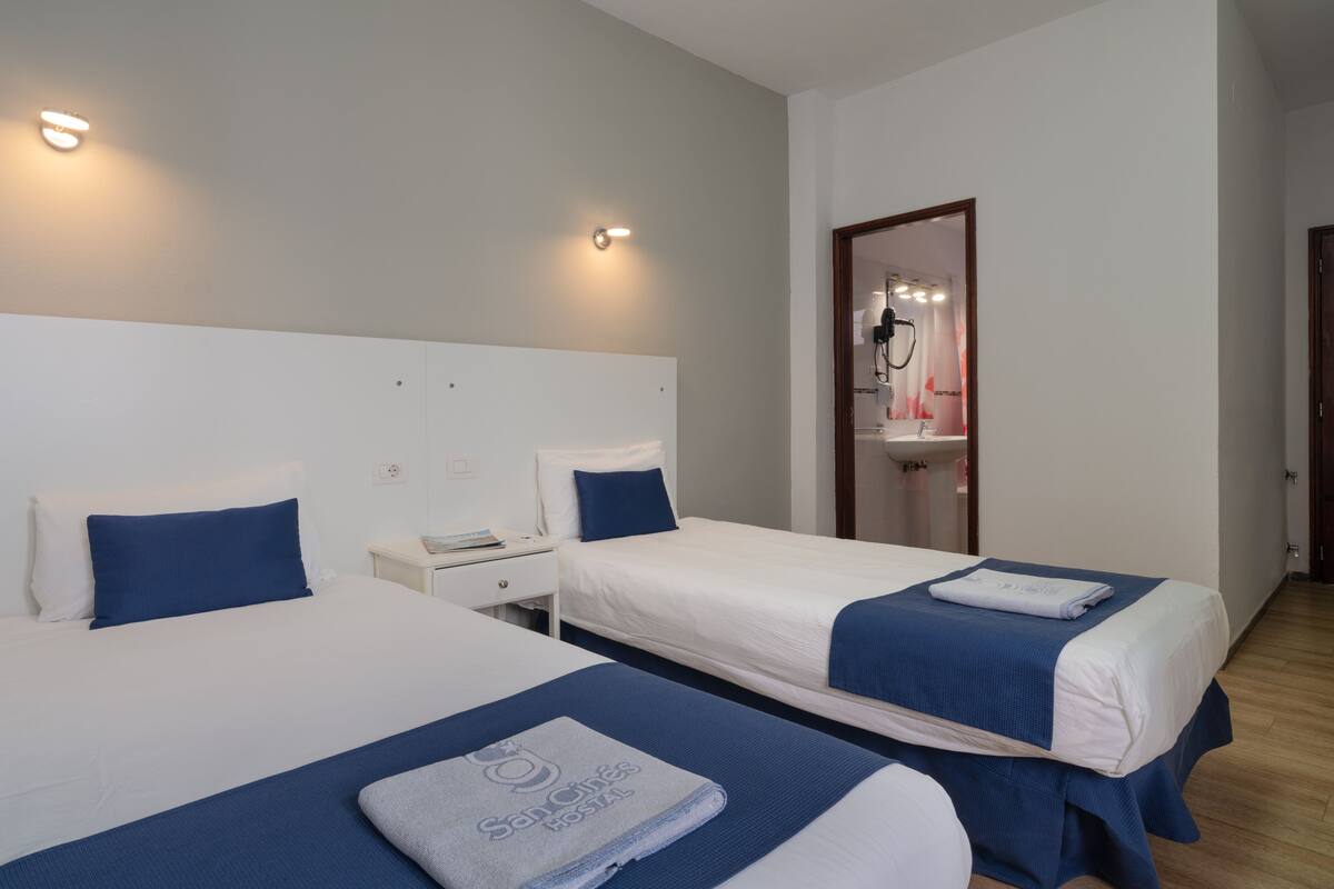 A hotel room with two beds, each with a blue bedspread and a white headboard. There is a nightstand with a phone and a small table with a lamp. A bathroom is visible through an open door.