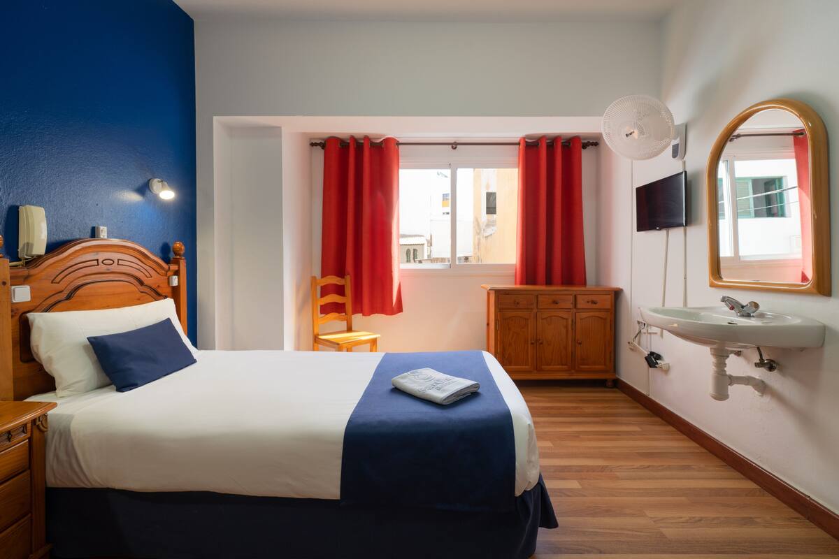 A hotel room with a bed, wooden nightstand, bedside table, a mirror, a sink, and a window with red curtains.