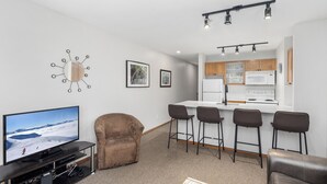 Un salon moderne avec un téléviseur à écran plat, un fauteuil marron et un coin bar avec des tabourets.