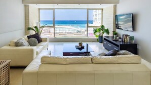 Ein modernes Wohnzimmer mit großem Fenster mit Blick auf Strand und Meer. Es gibt ein beiges Sofa, einen Couchtisch und einen an der Wand befestigten Fernseher.