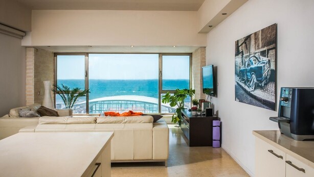 Ein modernes Wohnzimmer mit großem Fenster zum Meer, einem Sofa, einem Couchtisch und einem an der Wand befestigten Flachbildfernseher.