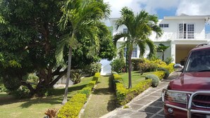 A well-maintained garden with palm trees, a statue, and a red car parked in front of a white building with a balcony. - 5 Bed Room Villa with Swimming pool  (Montego Bay)