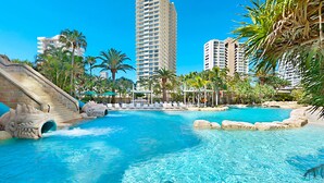 Una piscina de resort con una cascada, rodeada de palmeras y edificios modernos.