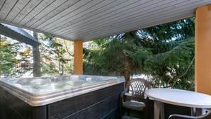 Un jacuzzi con revestimiento de madera, una mesa blanca y una silla en un espacio exterior cubierto con árboles al fondo.