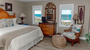 A bedroom with a bed, dresser, chair, and a view of the beach.