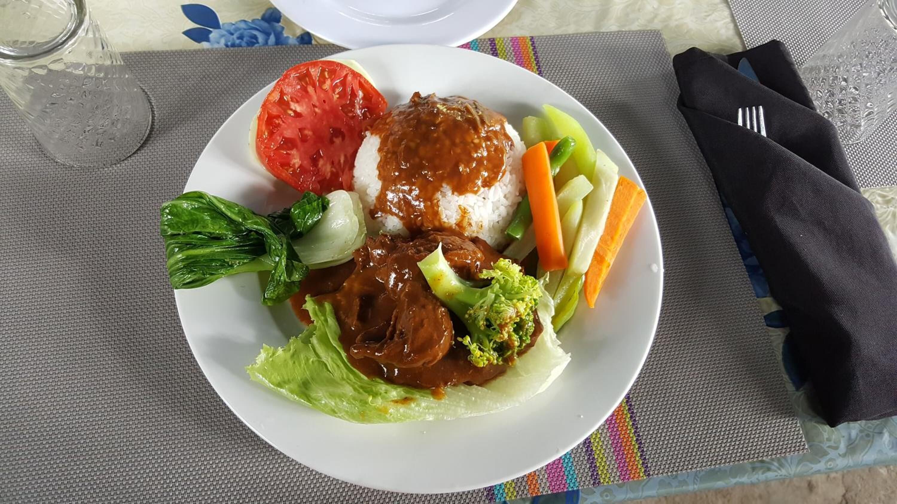 Ein Teller Essen mit Reis, Fleisch, Gemüse und einer Tomate auf einem Tisch.