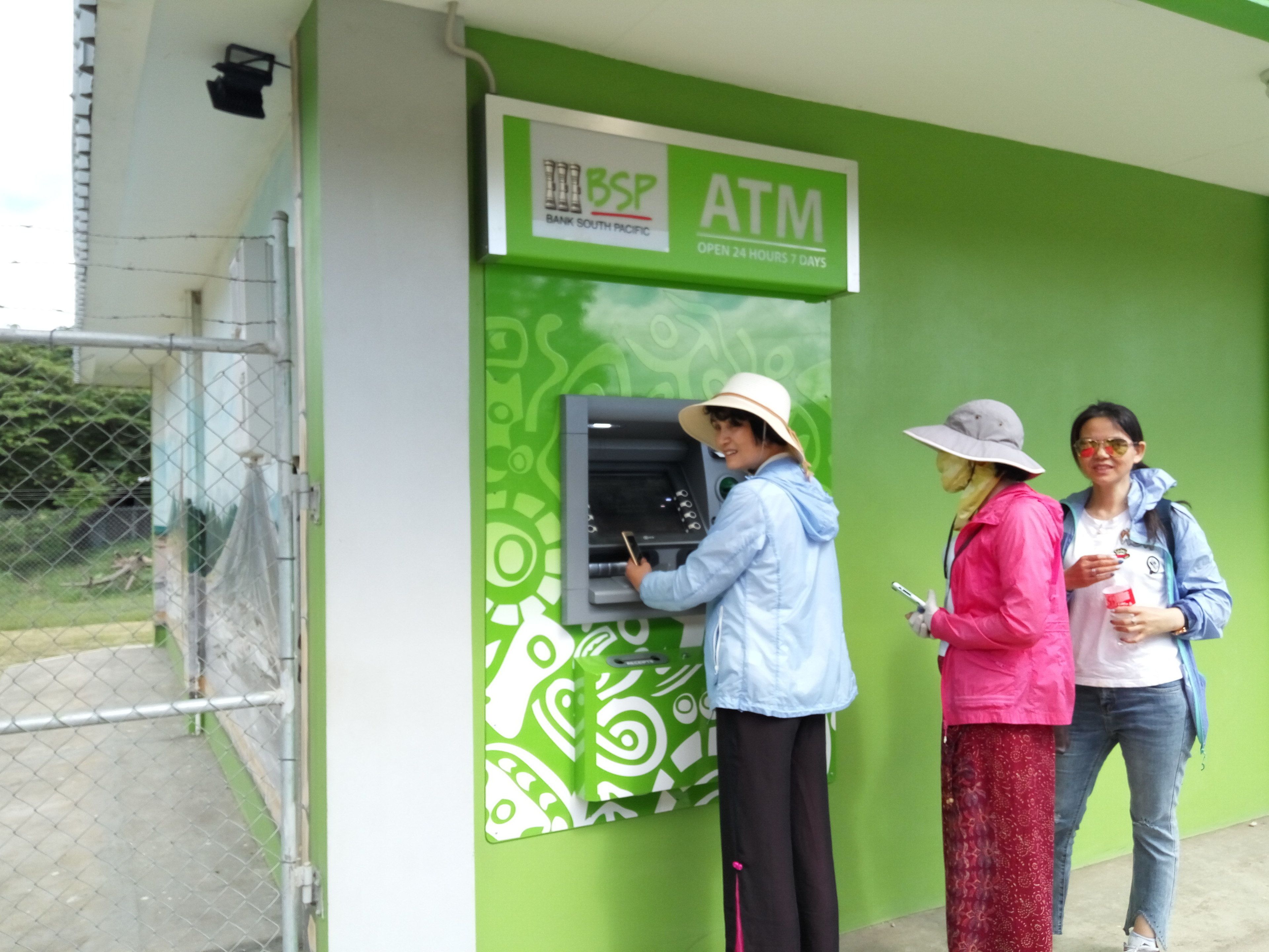 Un cajero automático con diseño verde y blanco, un cartel que indica que es un cajero automático de Bank South Pacific, y una cerca de malla metálica al fondo.