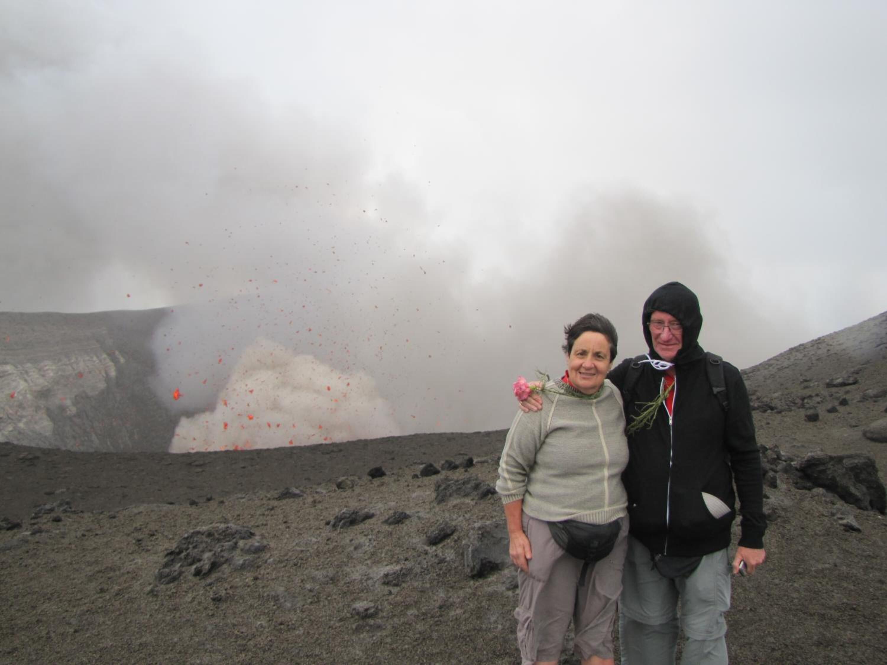 Two people standing on a volcanic landscape with a plume of ash and smoke in the background.