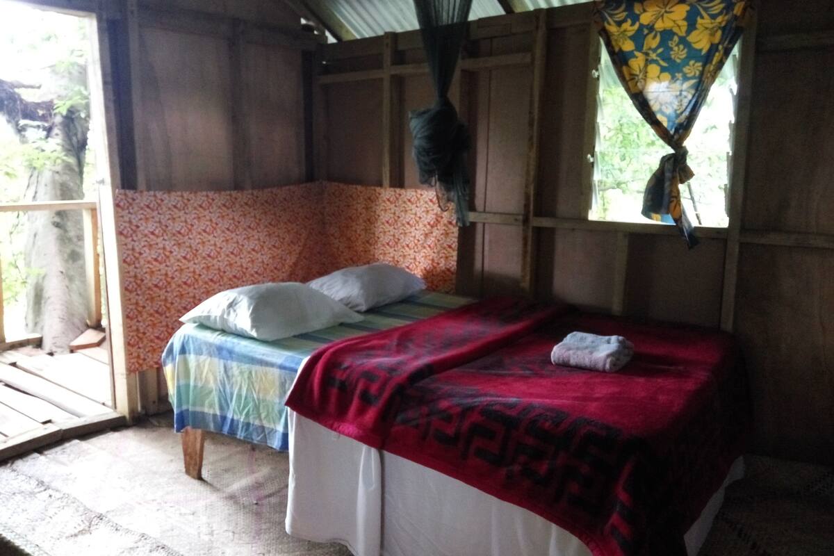 A single bed with a red blanket and pillows, a wooden floor, and a window with a curtain.