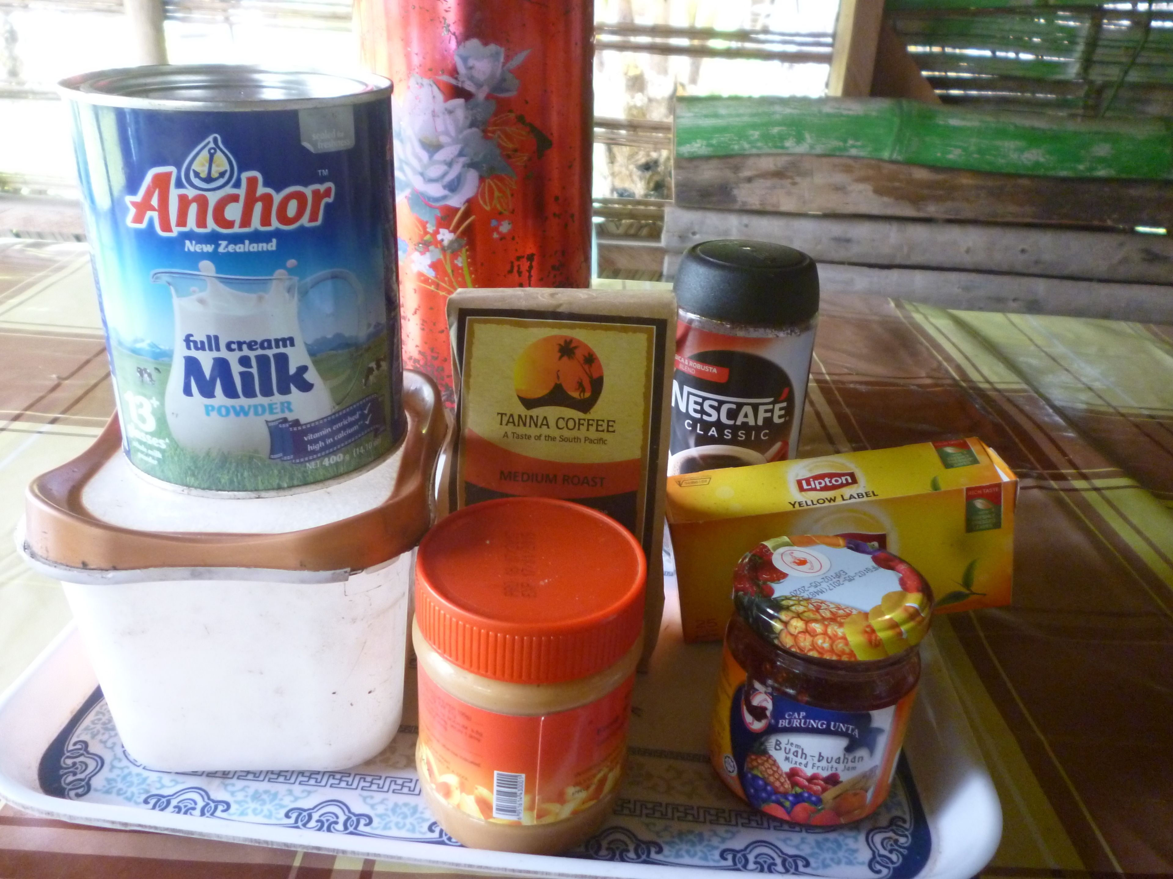Una bandeja con diversos alimentos, incluyendo una lata de leche entera Anchor, una lata de café Tanna, un tarro de té Lipton, un tarro de Cap Burung Unta y un tarro de frutas mixtas.