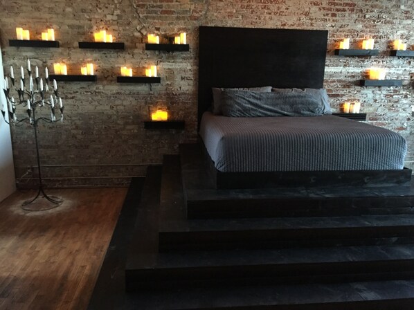 A modern bedroom with a brick wall, a bed with a dark headboard, and multiple wall-mounted shelves with candles.