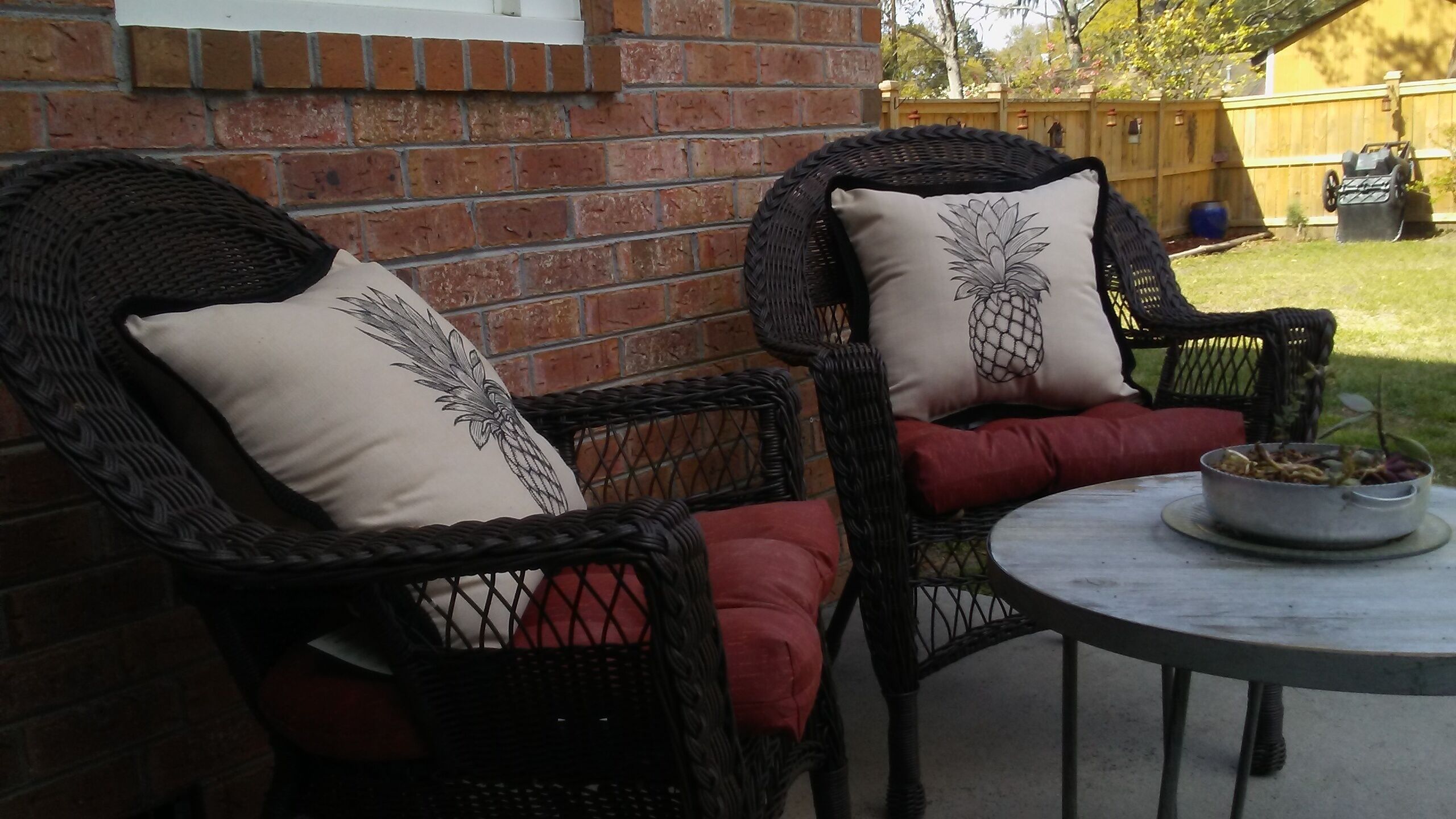 Two wicker chairs with cushioned seats and decorative pillows, a round metal table with a bowl of plants, and a brick wall in the background.