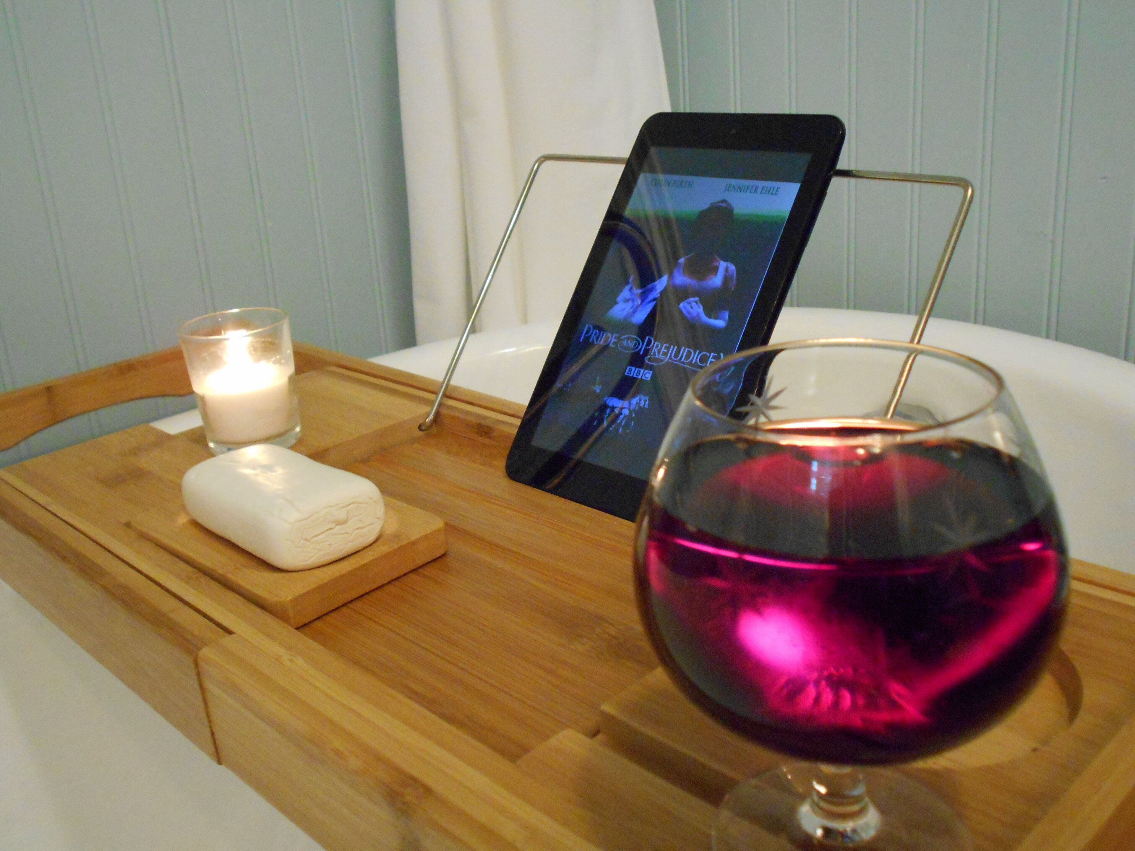 Un plateau en bois avec une bougie, un verre de vin rouge et une tablette affichant un programme télévisé.