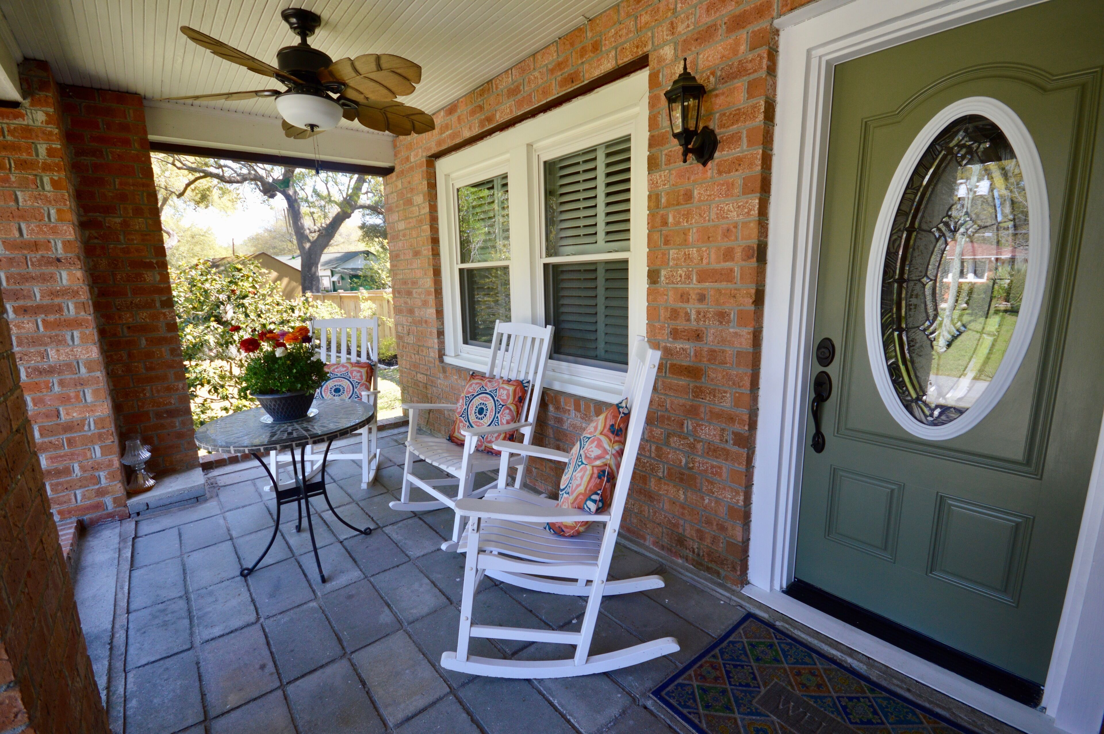 A porch with a brick wall, a green door, a white rocking chair, a table with a potted plant, and a ceiling fan.