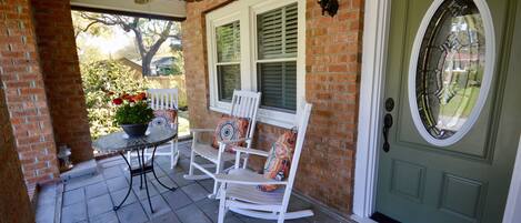 A porch with a brick wall, a green door, a white rocking chair, a table with a potted plant, and a ceiling fan.