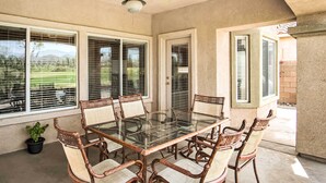 Un patio cubierto con una mesa de vidrio y sillas de mimbre, ventanas grandes con persianas y un ventilador de techo.