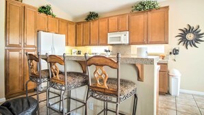 Una cocina con gabinetes de madera, un refrigerador blanco, un microondas y dos taburetes de bar con cojines estampados.