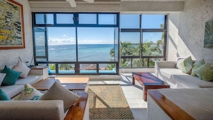 A spacious living room with large windows offering ocean views, a ceiling fan, and a cozy seating area with cushions and a coffee table. - Amazing View Oceanfront with 2 pools, Daily Housekeeping (Akumal)