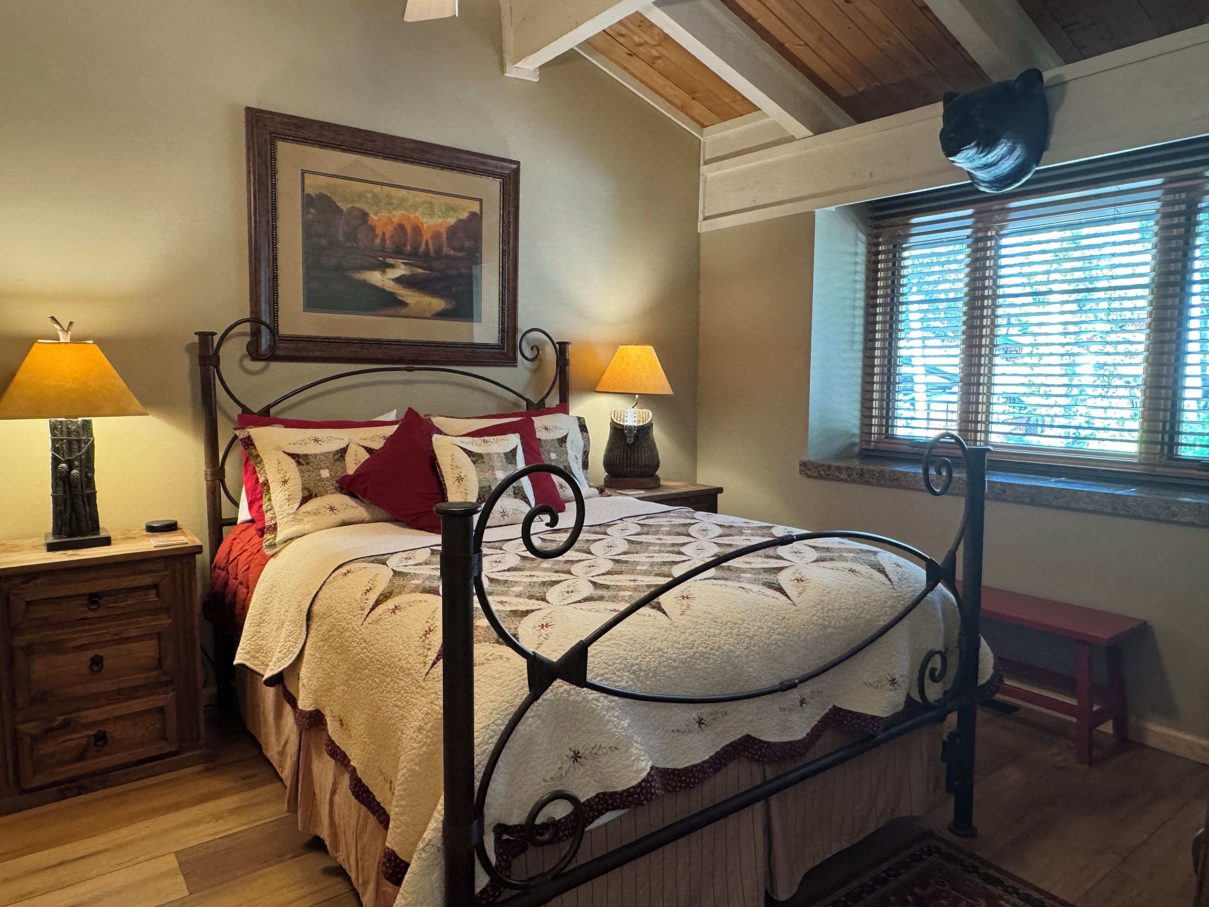 Une chambre avec un lit, deux tables de chevet, une lampe, un banc, une fenêtre avec des stores et une décoration représentant une tête d’ours au plafond.