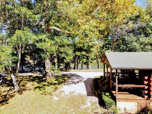 A wooden cabin with a porch, surrounded by trees and a dirt path leading to a lake.
