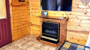 Interior acolhedor de uma cabana, com parede de madeira, lareira, um certificado emoldurado e televisão montada acima.