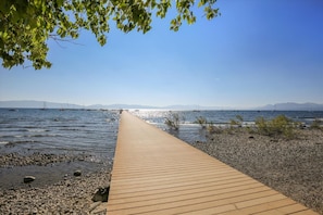 A wooden boardwalk leading to a rocky shoreline with boats in the water and mountains in the distance.