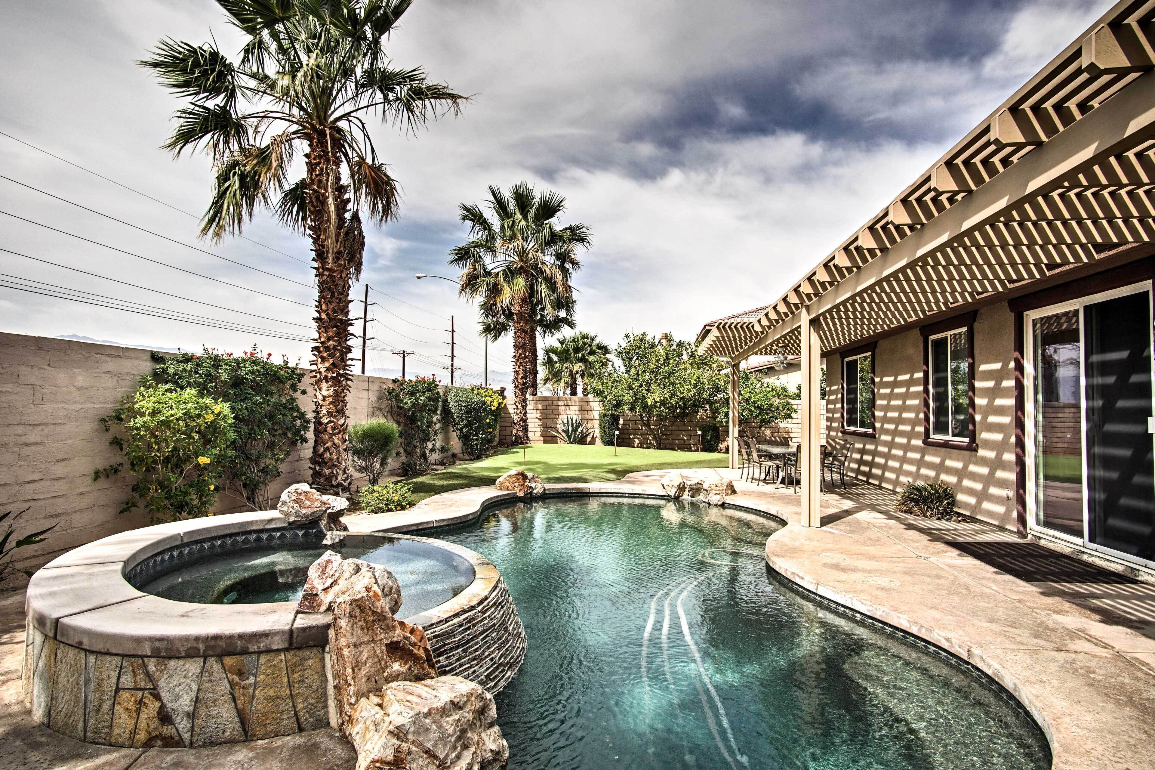 A swimming pool with a hot tub, surrounded by palm trees and a patio area.
