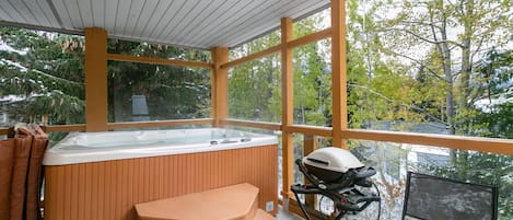 A hot tub with wooden seating and a grill, surrounded by large windows offering a view of trees.