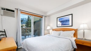 Ein Hotelzimmer mit Bett, Schreibtisch, Stuhl, Nachttisch und Fenster mit Blick auf Bäume und Berge.