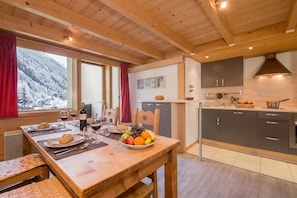 A wooden interior with a dining table set for a meal, a kitchen area with cabinets and a stove, and a window with a view of snow-covered trees. - Grand Roc A218 150m from Slopes, View & Parking, Argentière (Chamonix), France (Argentière (Chamonix))