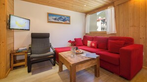 A wooden-paneled room with a red sofa, a grey armchair, a wooden coffee table, a TV on the wall, and a window with a view of buildings. - Grand Roc A218 150m from Slopes, View & Parking, Argentière (Chamonix), France (Argentière (Chamonix))