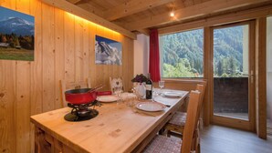 A wooden dining table with chairs, a red pot, wine glasses, and a bottle. There are two paintings on the wall and a window with a red curtain. - Grand Roc A218 150m from Slopes, View & Parking, Argentière (Chamonix), France (Argentière (Chamonix))