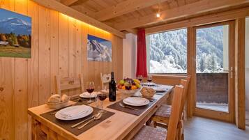 Une table de salle à manger en bois, mise avec des assiettes, des verres et une bouteille, avec vue sur la montagne par la fenêtre.
