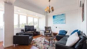 A modern living room with a flat-screen TV, comfortable seating, a coffee table, and a large window offering a view of trees. - Cozyplace 23 in Lisbon - 1 bedroom appartment (Lisboa)