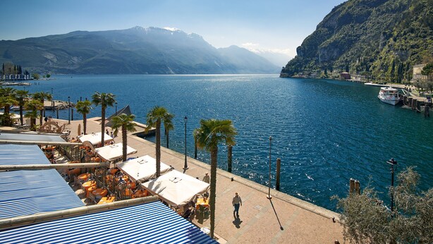 A waterfront hotel with outdoor seating, palm trees, and a view of a lake and mountains. - Appartamento Villa Lina with Lake View, Riva del Garda, Italy (Riva del Garda)