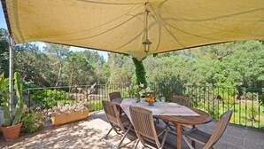 Een terras buiten met een tafel en stoelen, omringd door een metalen hek en een grote gele luifel.
