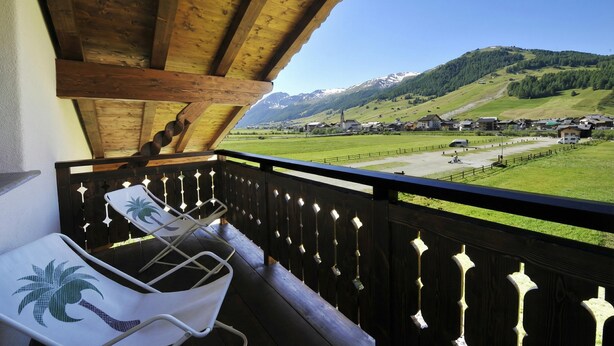 A balcony with a wooden railing, a white chair with a palm tree design, and a view of a village with mountains in the background. - Arcobaleno, Livigno, Italy (Livigno)