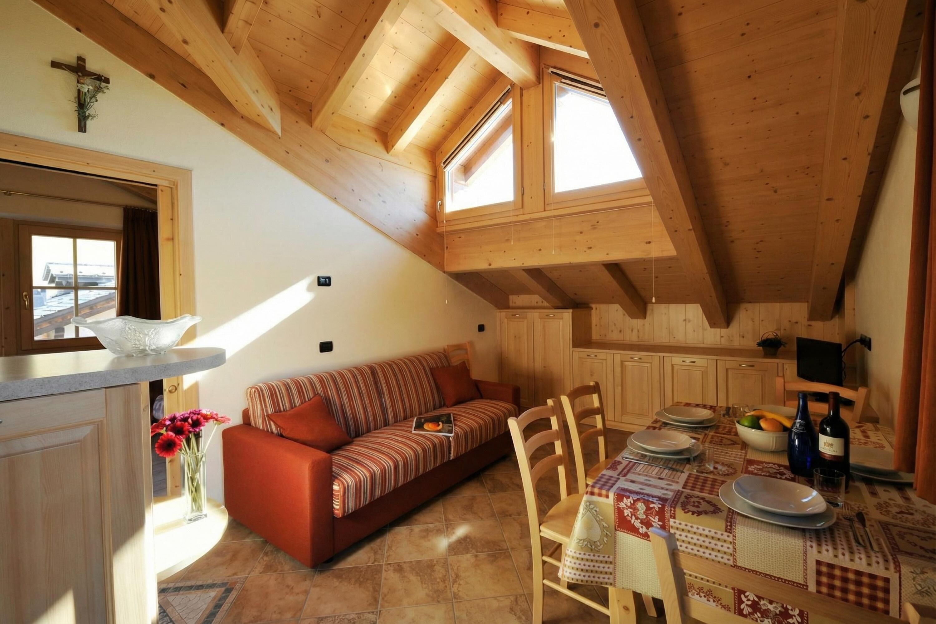 A cozy loft-style room with a wooden ceiling, a sofa, a dining table set for two, and a kitchen area.
