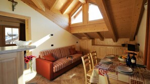 A cozy loft-style room with a wooden ceiling, a sofa, a dining table set for two, and a kitchen area. - Arcobaleno, Livigno, Italy (Livigno)