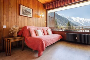 Une pièce en bois avec un lit, une table sur laquelle se trouve un vase, une lampe fixée au mur et une fenêtre donnant sur la montagne.