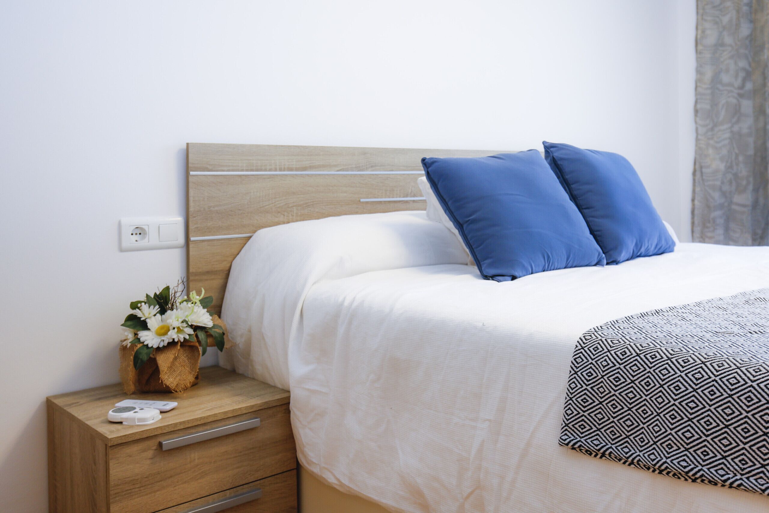 Un letto ben rifatto con poggiatesta in legno, un comodino con un cesto di fiori e una presa elettrica sulla parete.