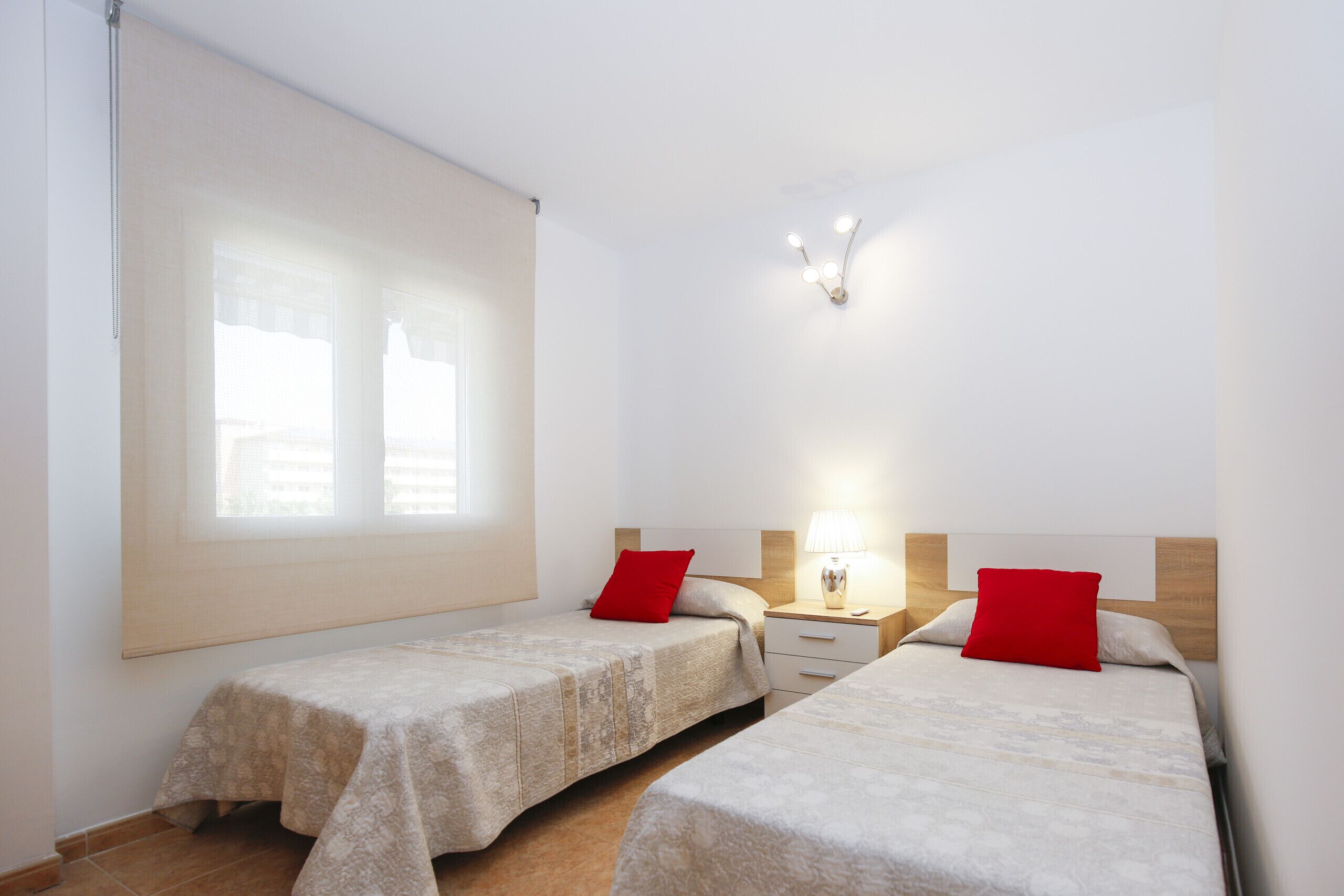 A hotel room with two beds, a nightstand with a lamp, a window with blinds, and a wall-mounted light fixture.