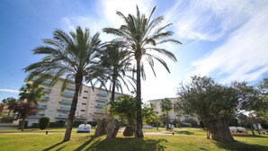A landscaped area with palm trees, a grassy lawn, and a building with multiple balconies in the background. - LOS JUNCOS HomeStay by Turismar (La Pineda)