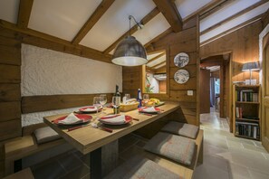 Una mesa de comedor de madera dispuesta con platos, vasos y una cesta de frutas. Alrededor de la mesa hay bancos acolchados.