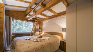 Ein gemütliches Schlafzimmer mit Holzbalkendecke, einem großen Bett mit gemustertem Überwurf, einem Nachttisch mit Lampe und einem Fenster mit Vorhängen.