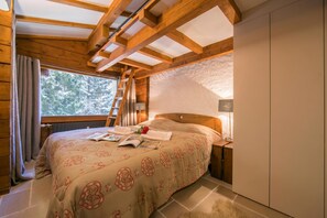 Une chambre confortable avec un plafond en bois, une grande fenêtre, un lit recouvert d’une couvre-lit à motifs et une table de chevet avec une lampe.