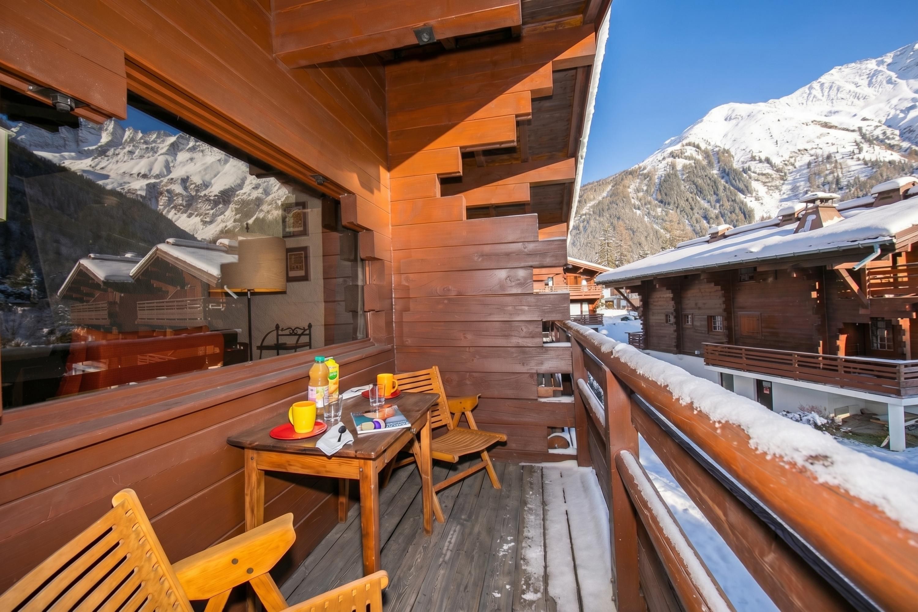 Een houten terras met een tafel en stoelen, uitkijkend op een besneeuwd berglandschap.