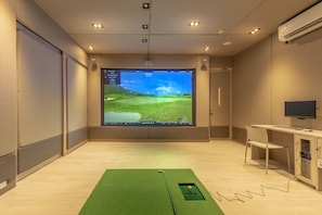 Una habitación con una pantalla grande mostrando un juego de simulación de golf, un tapete de práctica verde, un escritorio con una silla y un equipo de aire acondicionado.
